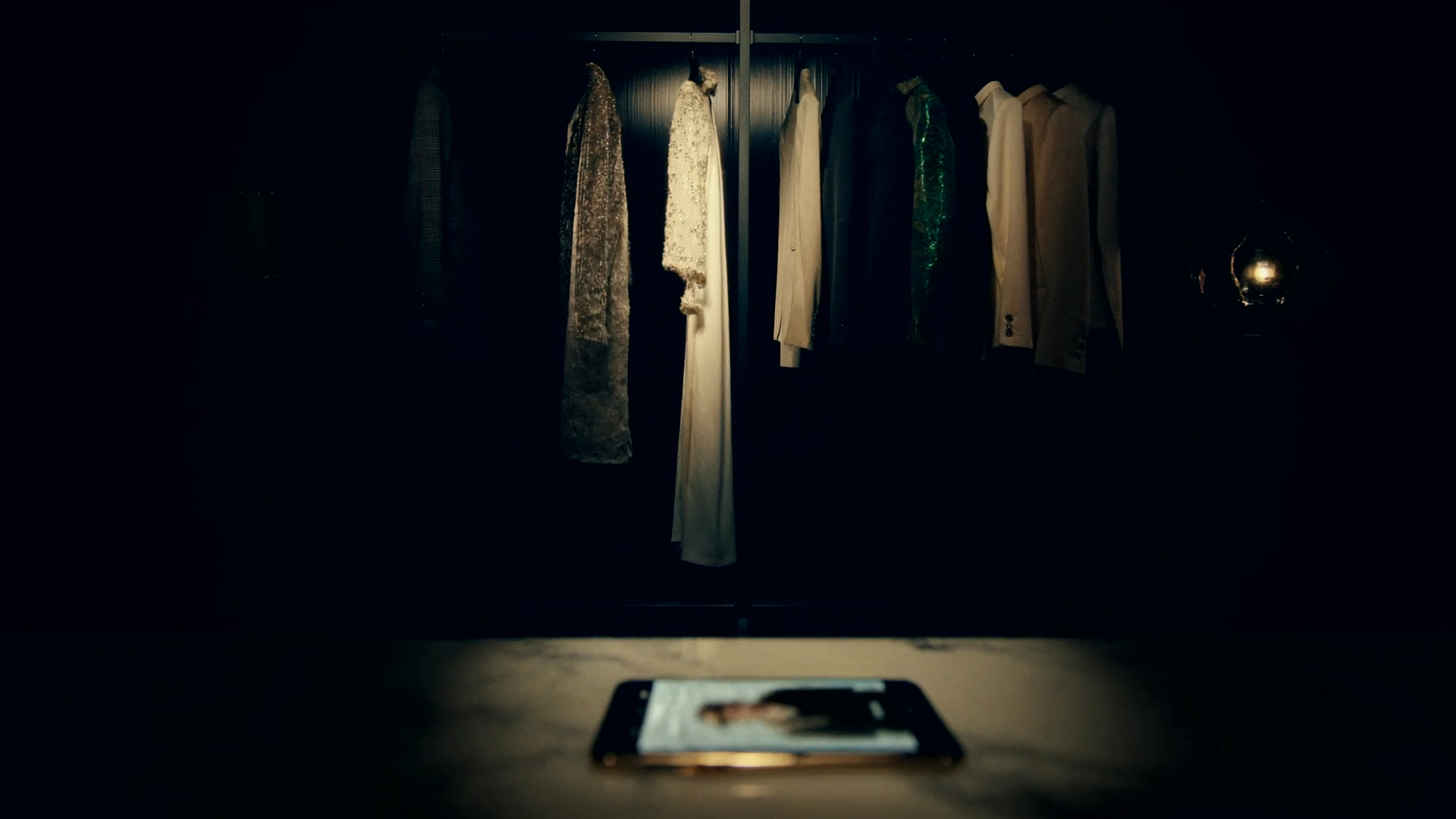 Perchero de prendas premium iluminado en penumbra cálida junto a un teléfono que muestra una fotografía de una prenda